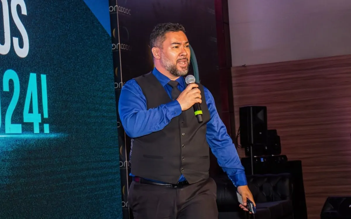 Clayton no palco como mestre de cerimônias, de colete e camisa azul, falando ao microfone.