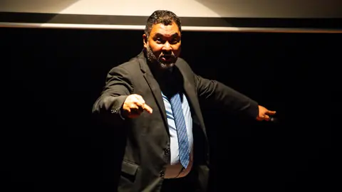 Clayton Yaso no palco escuro, apontando com os dois braços, em pose de direção de cena.