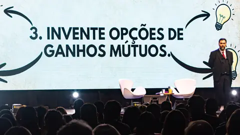 Vista da plateia: o Clayton Yaso no palco ao lado do slide “3. Invente opções de ganhos mútuos”, com uma lâmpada desenhada.