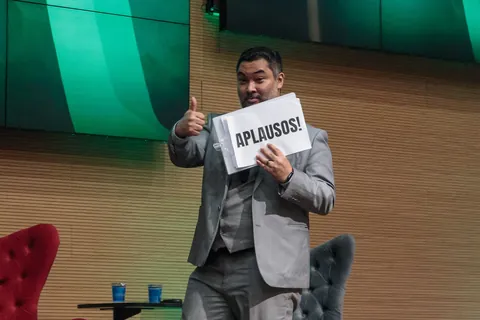 Clayton Yaso no palco do CONAPREF segurando a placa “APLAUSOS!” e fazendo sinal de positivo para a plateia.