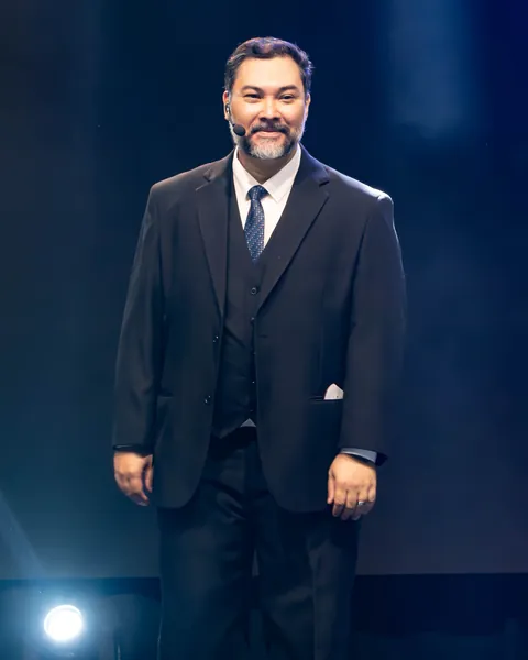 Clayton Yaso no palco de terno escuro e microfone de cabeça, sorrindo para a plateia sob a luz de um refletor.