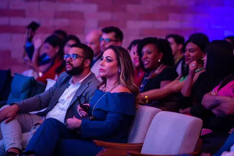 Participantes na plateia sob luz rosa, acompanhando a palestra com atenção.